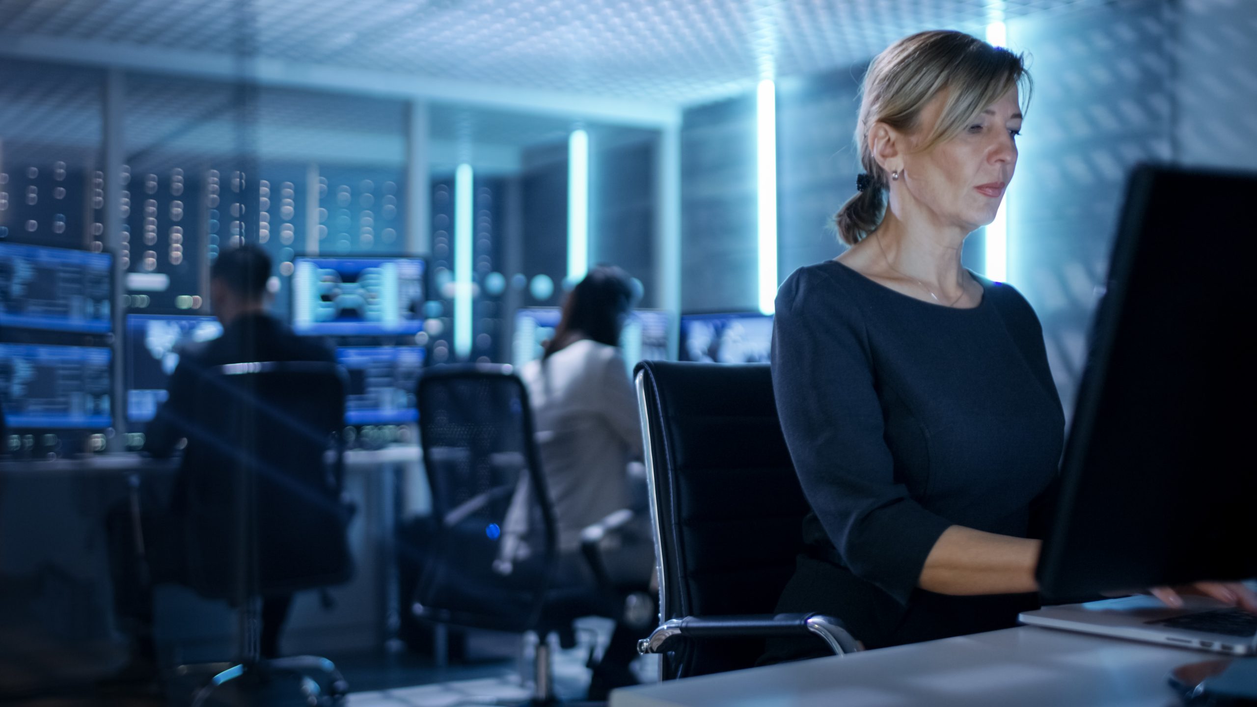 Female IT Engineer Works on Her Desktop Computer in Government Surveillance Agency. In the Background People at Their Workstations with Multiple Screens Showing Graphics.